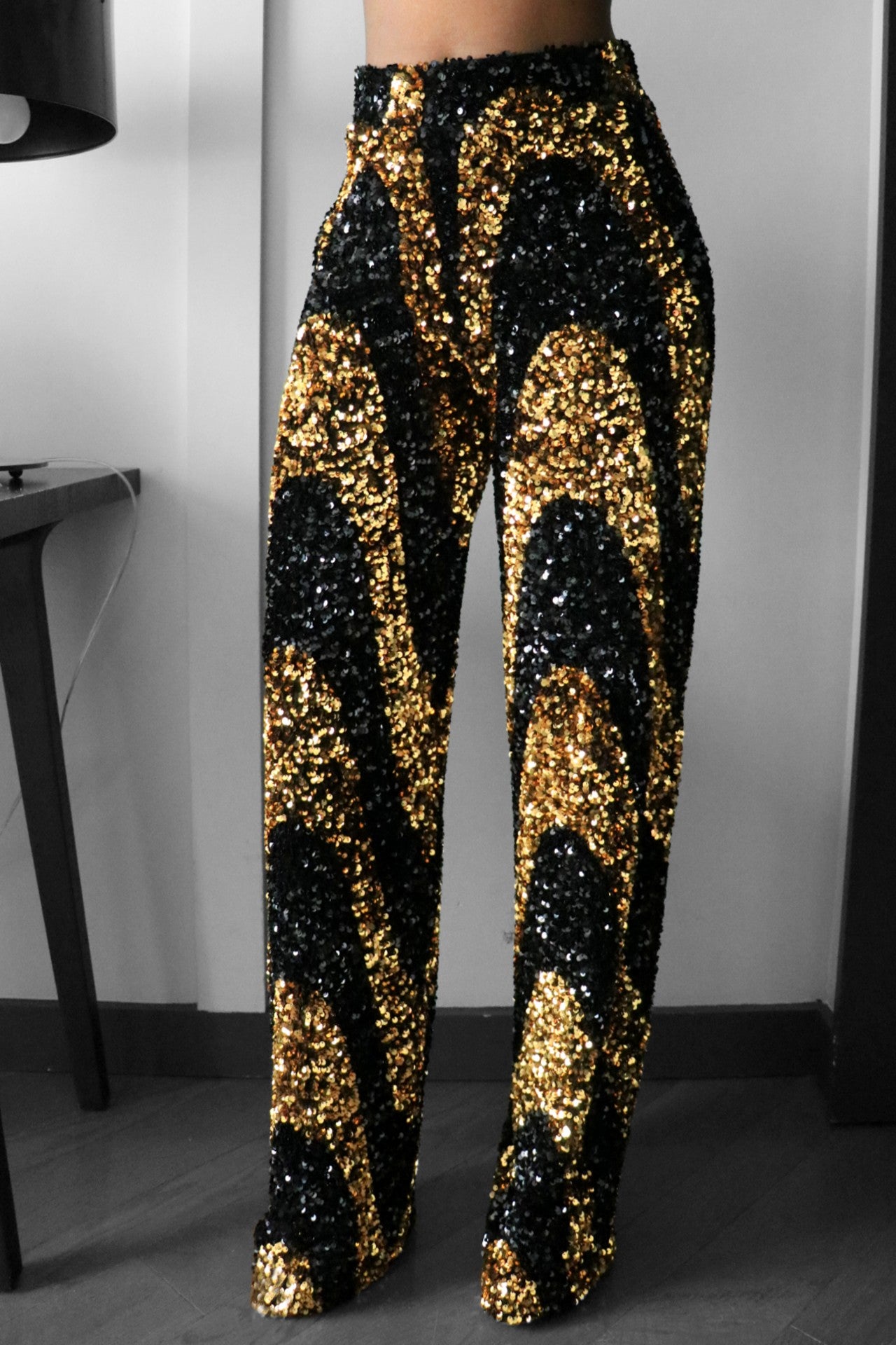 A pair of black and gold sequin wide leg pants with an elastic waist, shown in a reflective indoor setting.