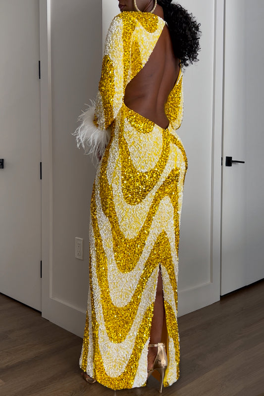 A woman shown from the back, wearing a floor-length sequined gown with a large open back, long sleeve, and a high back slit. The dress has a bold wavy pattern in metallic gold and white.