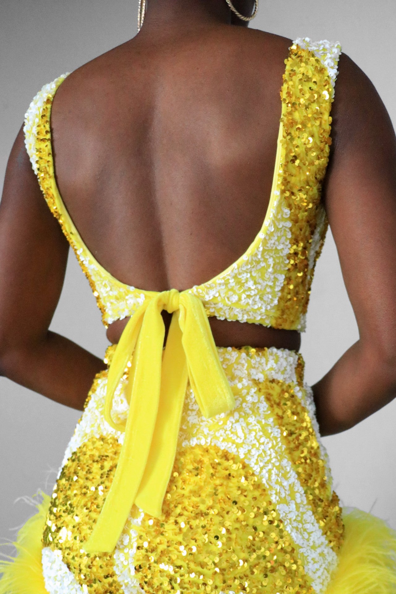 Full back view of a model wearing a yellow and white sequin swirl square neck crop top. Back is a deep scoop neckline that has a velvet ribbon tie.