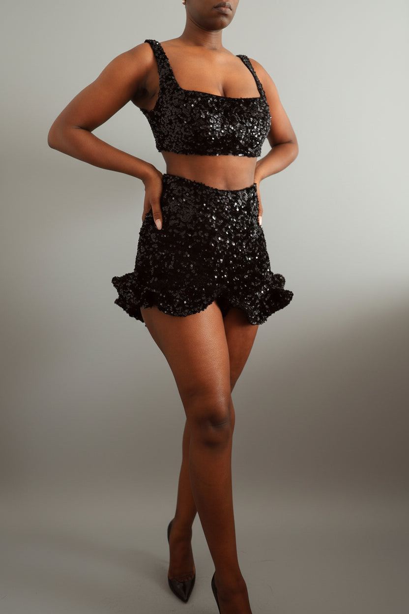 Model wearing a black sequin square neck bralette with matching sequin ruffle shorts against a plain wall.