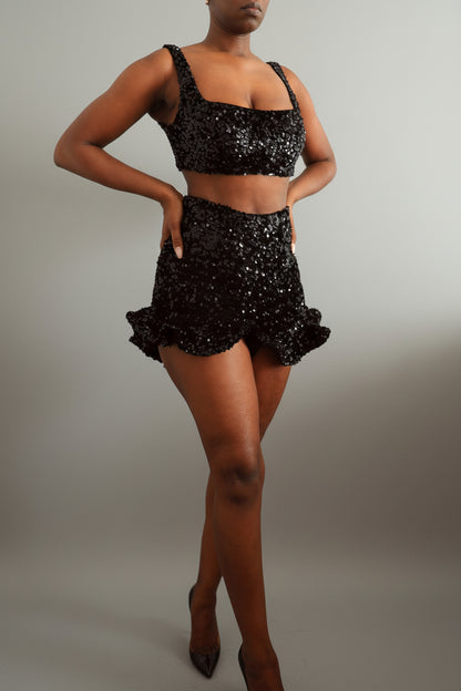 Model wearing a black sequin square neck bralette with matching sequin ruffle shorts against a plain wall.