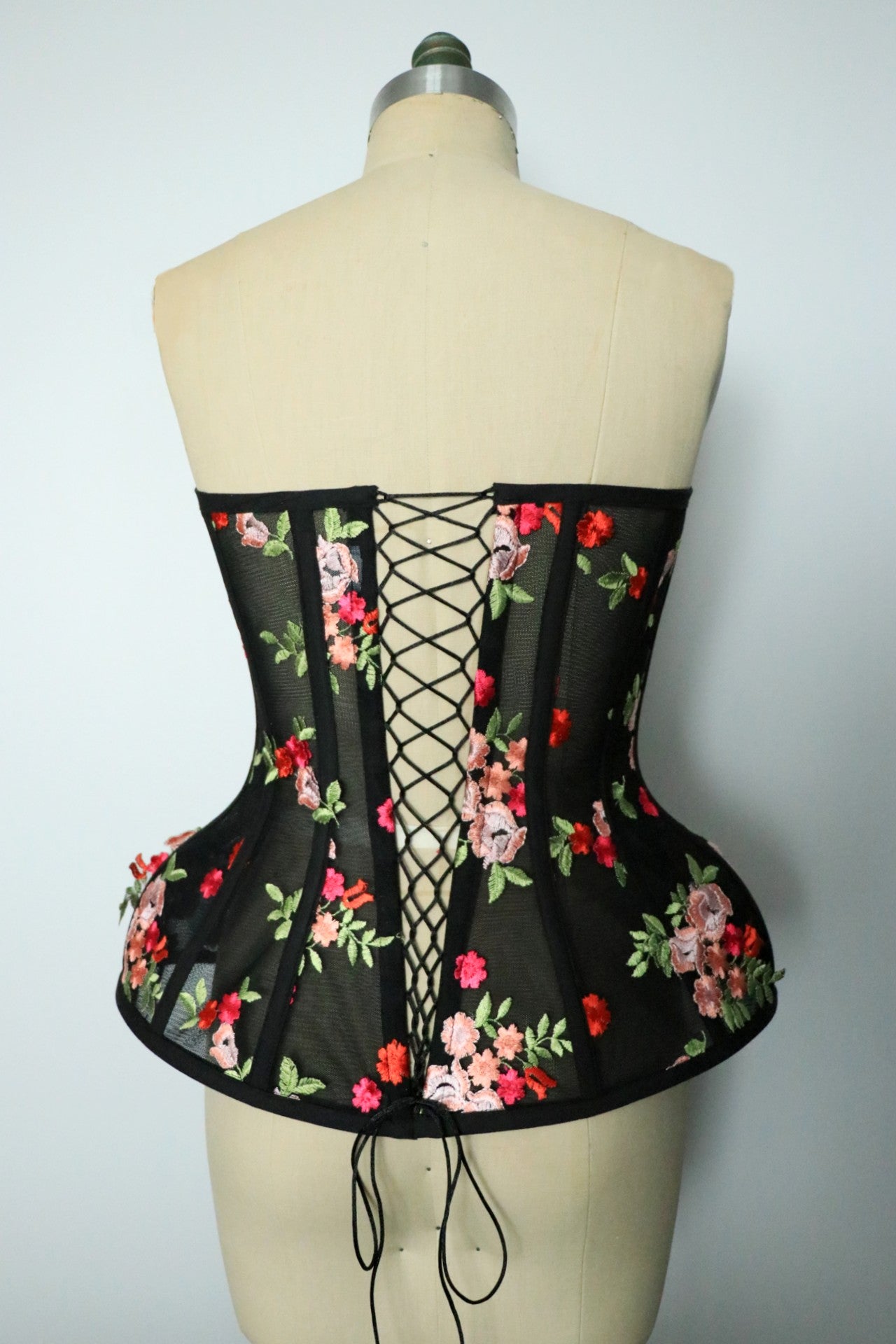 Back view of a black floral corset bustier is displayed on a beige dress form mannequin, featuring sheer mesh panels, a lace-up back, and red, pink, and green floral embroidery.