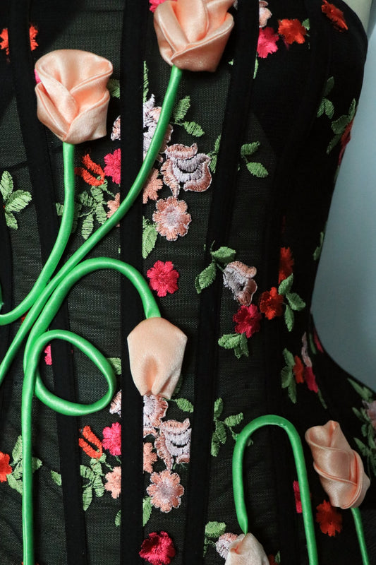 Close-up of a black, floral embroidered corset featuring peach-silk fabric roses with bright green stems.