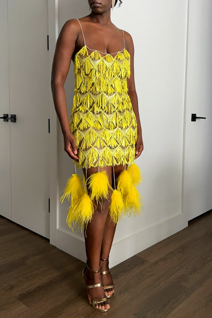 A woman modeling a bright yellow sequin-fringe dress with a feathered-tassel hem and thin rhinestone shoulder straps, standing in an indoor room with white paneled walls and wood flooring.
