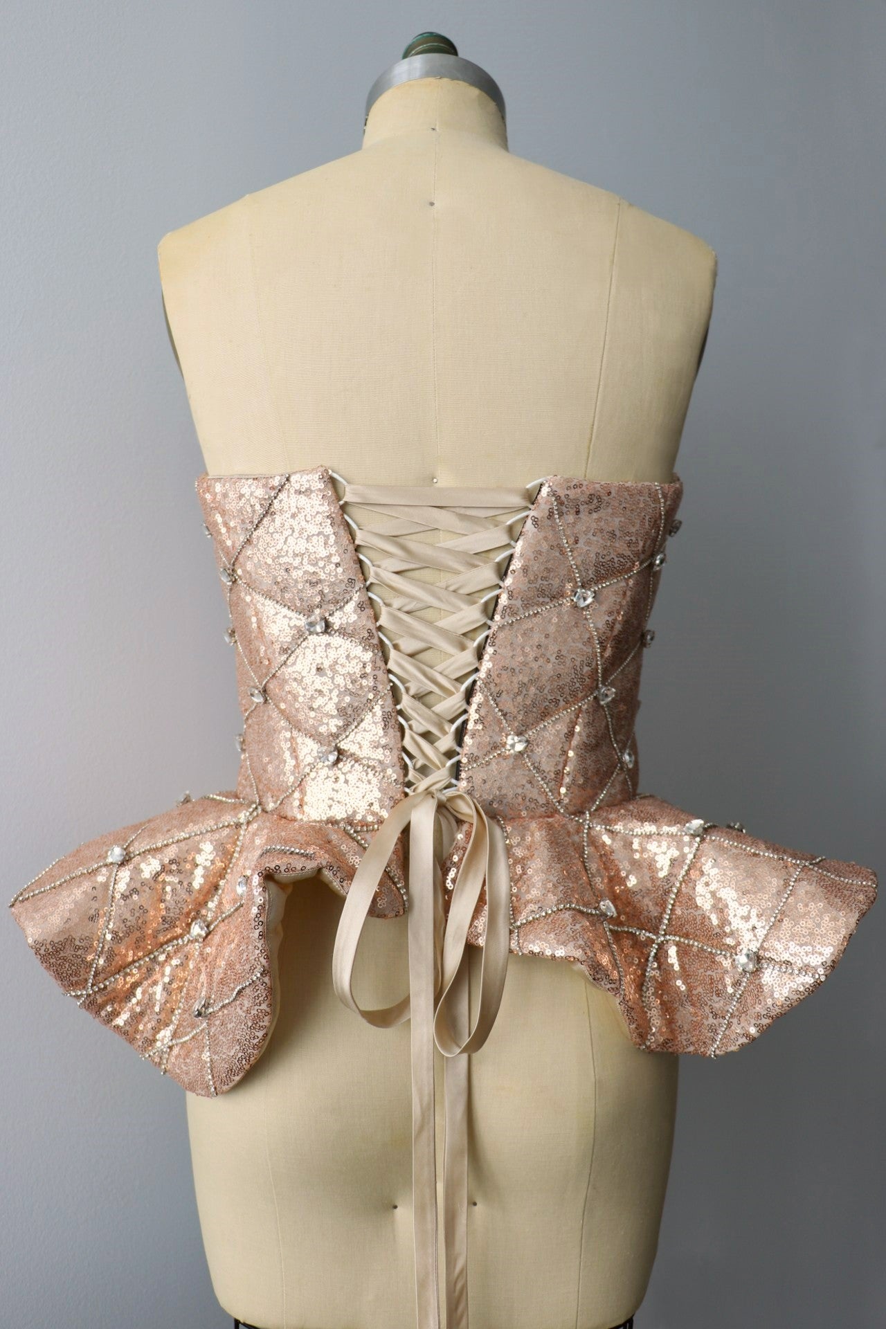 Back view of a rose gold quilted sequin peplum heart rhinestone corset with satin back lace up closure on a dress-form against a gray background 