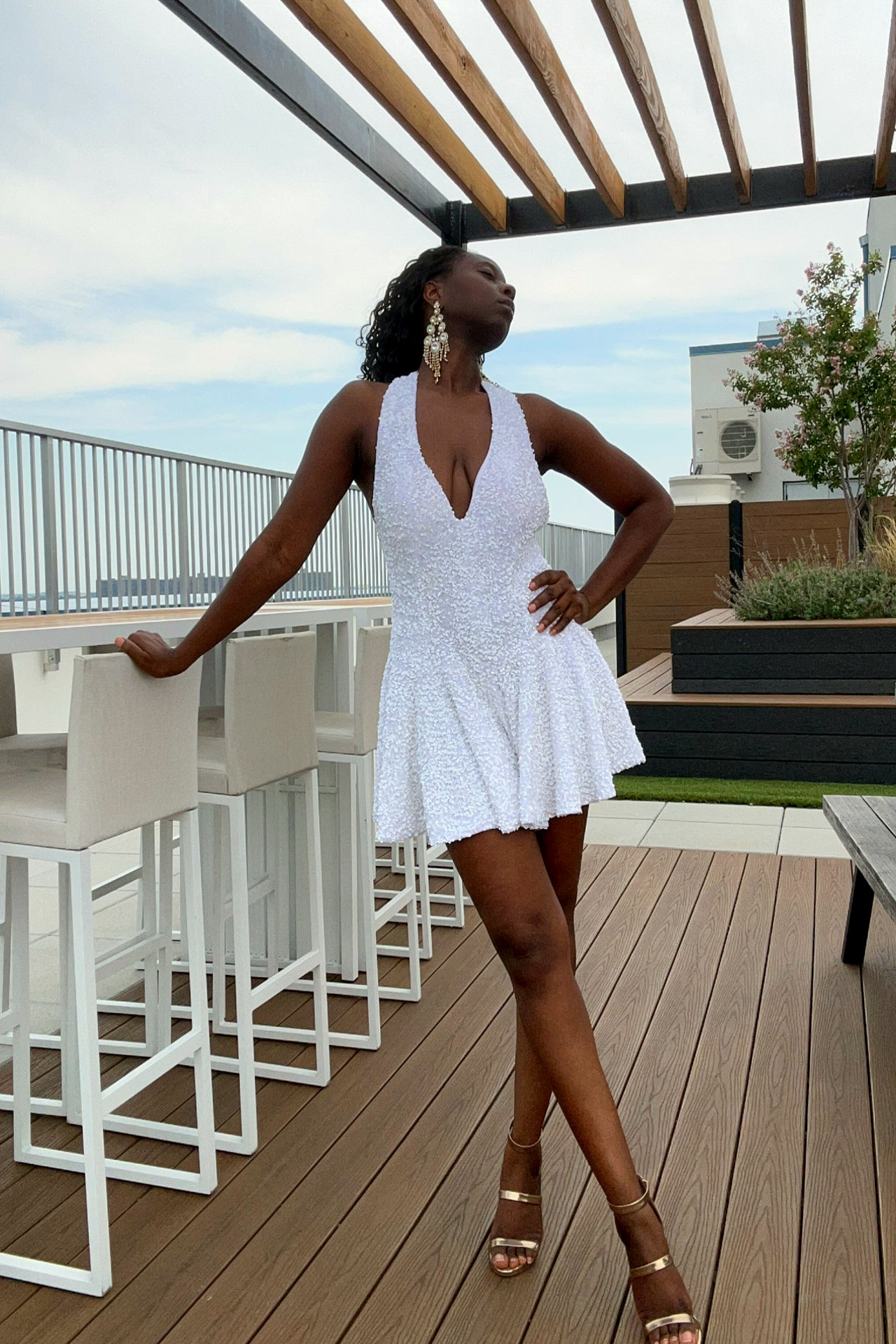 A woman posing on a rooftop terrace wearing a white sequin mini romper dress with a plunging neckline and gold high-heeled sandals.