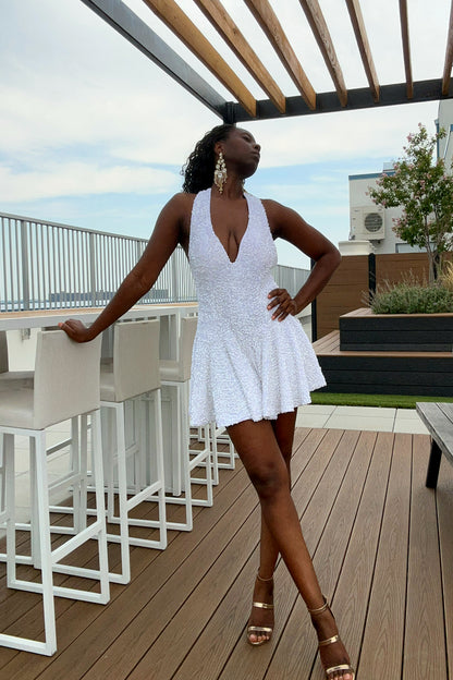 A woman posing on a rooftop terrace wearing a white sequin mini romper dress with a plunging neckline and gold high-heeled sandals.