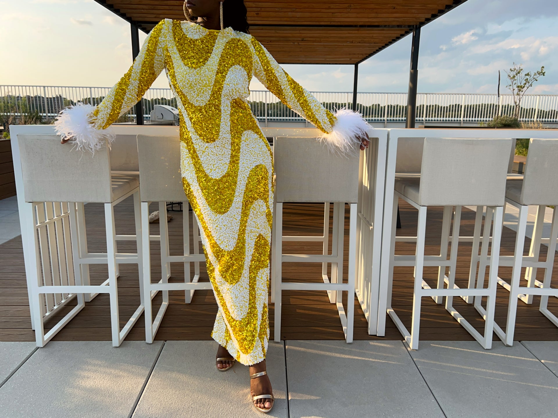 A woman stands on an outdoor patio wearing a long-sleeve maxi dress covered in yellow and white wavy sequins, with white feathered cuffs and gold strappy heels.