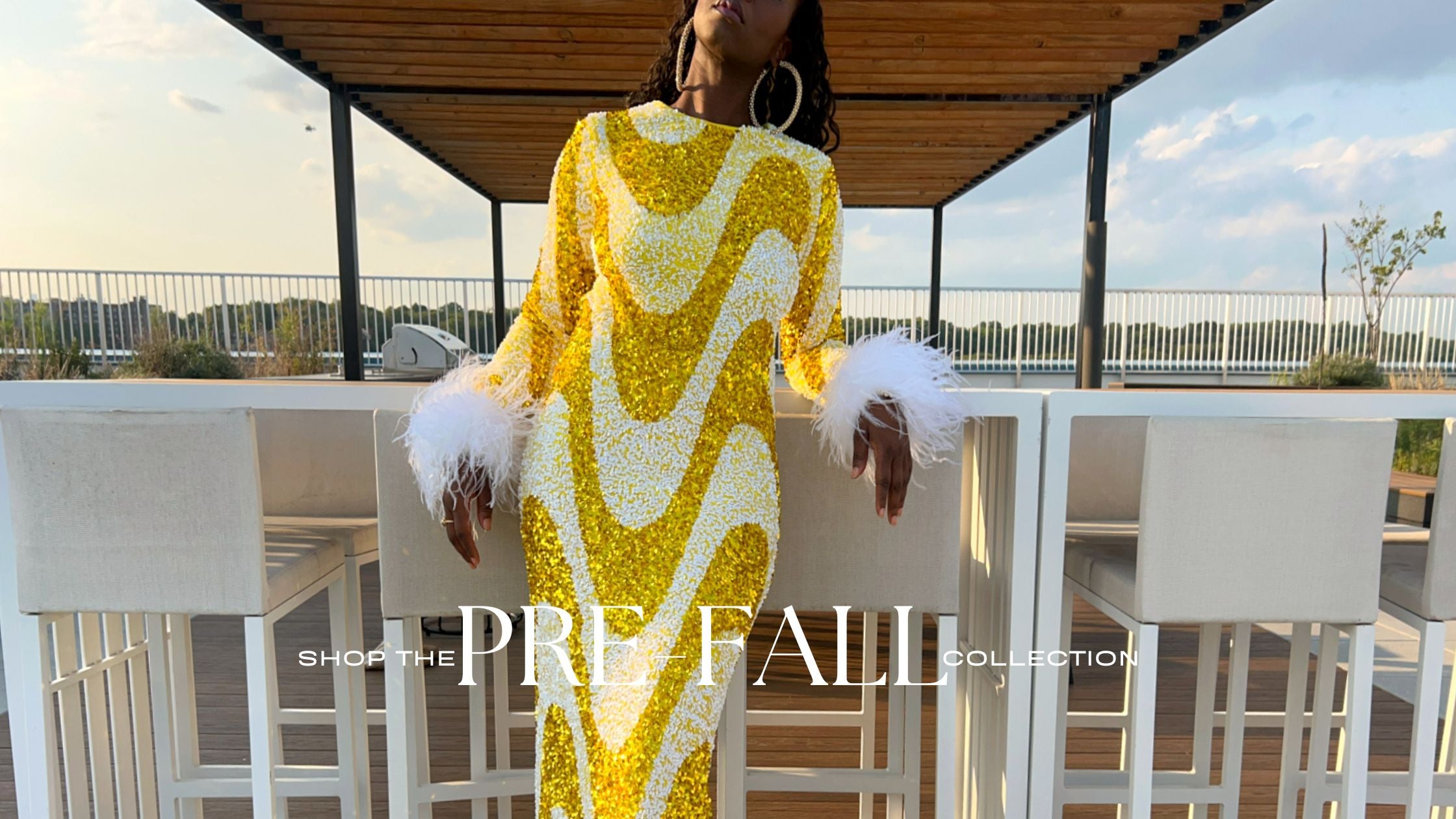 A model wearing a long-sleeved yellow and white sequined wavy pattern dress with white feathered cuffs, standing on an outdoor patio with white bar stools under a wooden roof.