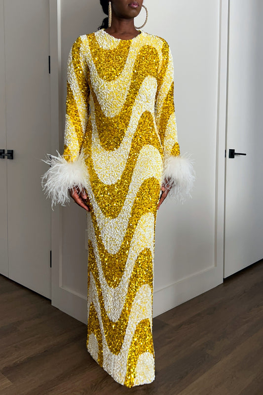 A woman wearing a long-sleeve maxi dress covered in gold and white sequins in a wavy pattern, with white feather trim at the cuffs, standing indoors against white paneled doors.