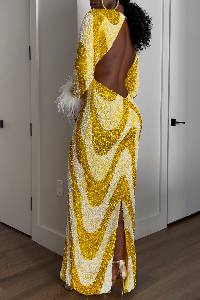 A woman shown from the back, wearing a floor-length sequined gown with a large open back, long sleeve, and a high back slit. The dress has a bold wavy pattern in metallic gold and white.