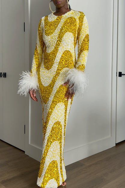 A woman wearing a floor-length sequined gown with a bold wavy pattern in gold and white sequins, featuring white feathered cuffs, standing in an indoor room with white doors and wood flooring.