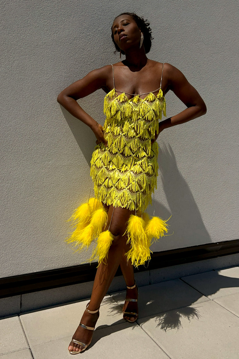 A woman models a bright yellow fringed Sequin dress with rhinestone feather tassels at the hem, standing against a light gray wall.