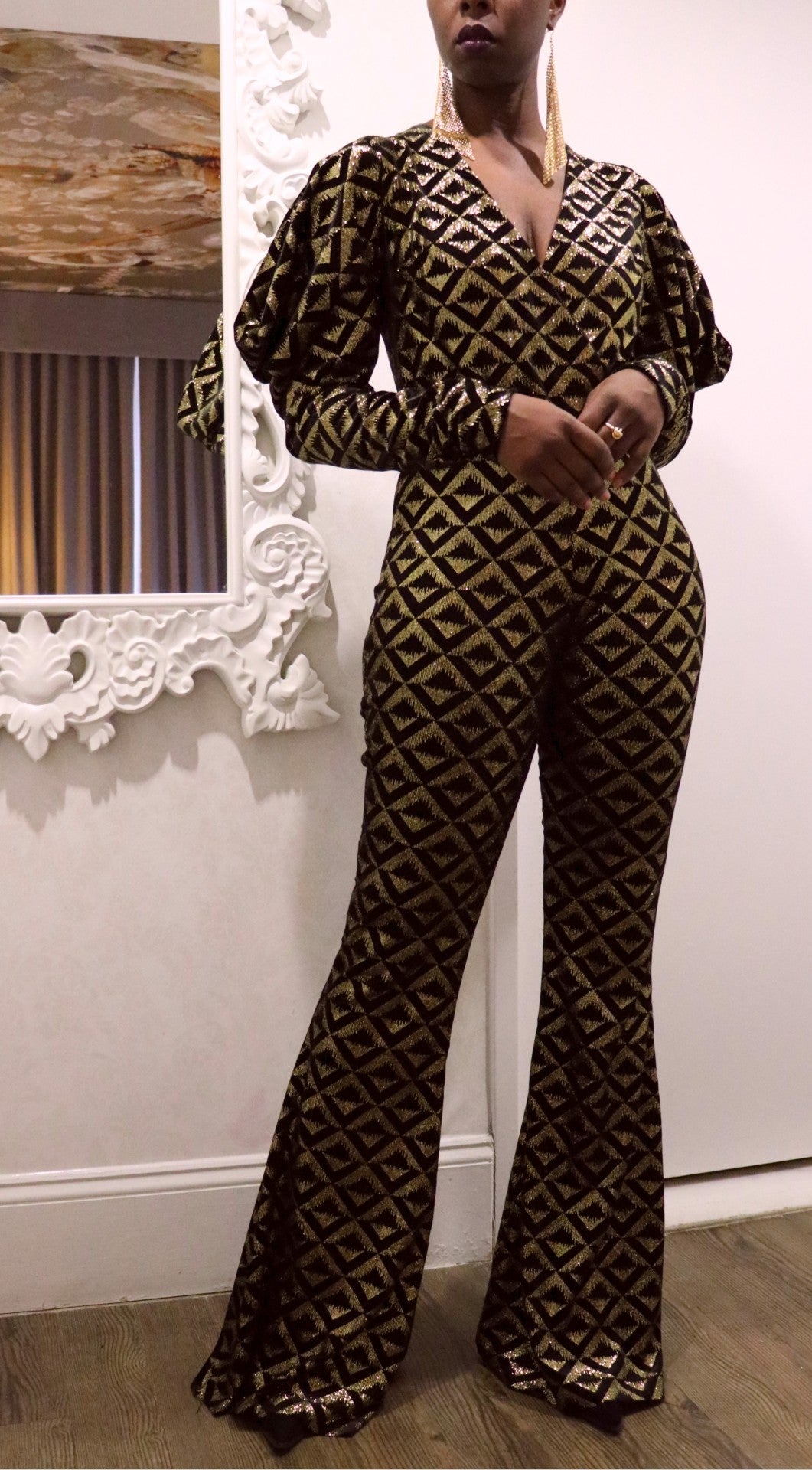 The model is wearing a black and gold geometric-patterned jumpsuit, standing against a white wall.