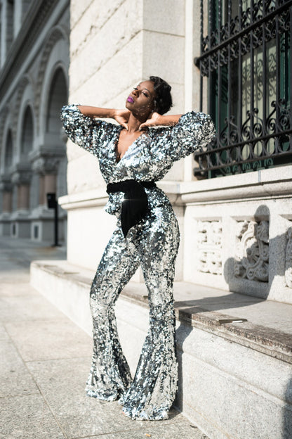 A model wearing a silver sequin jumpsuit is standing in front of a building.