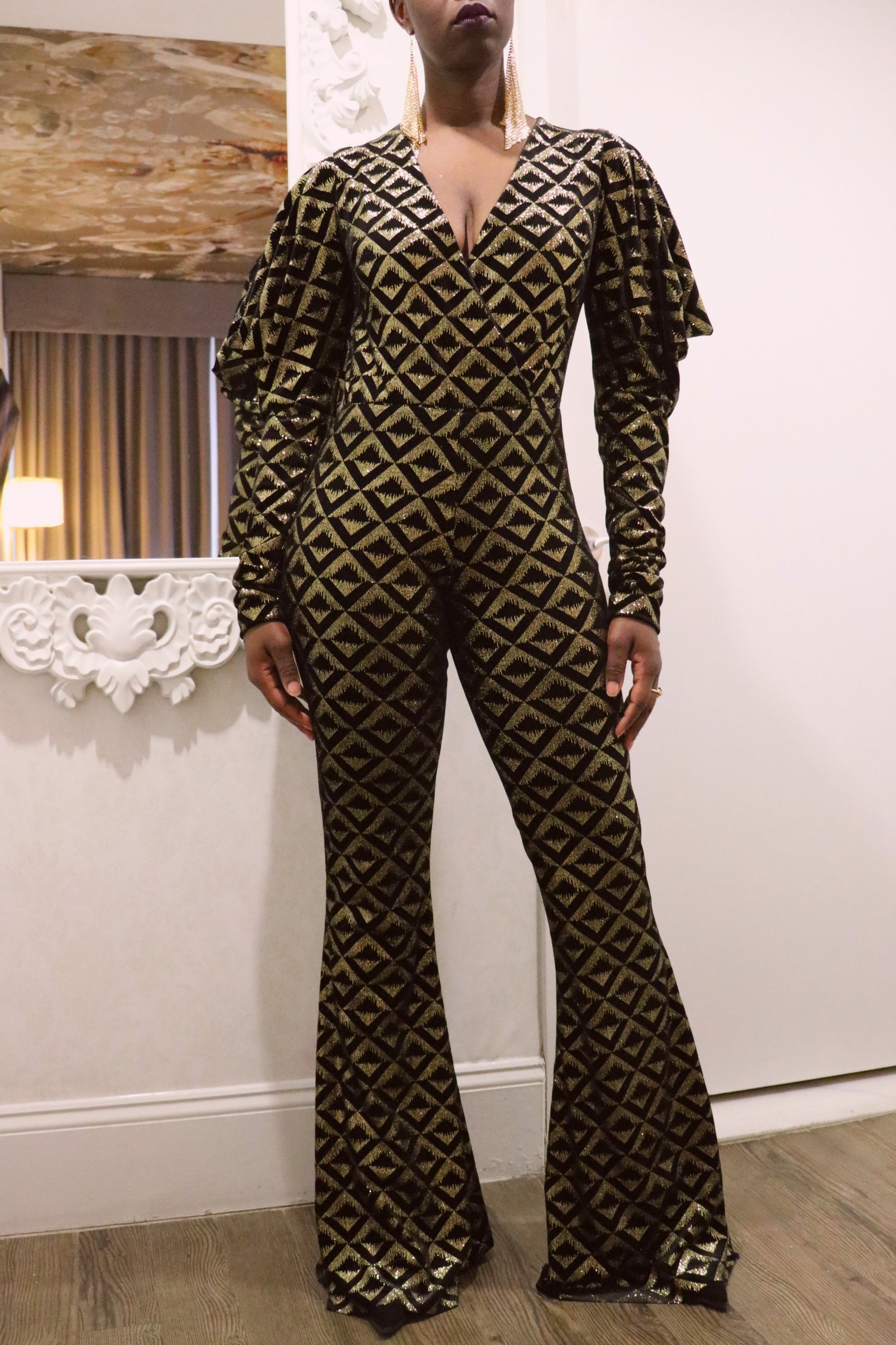 Model wearing a black and gold geometric-patterned jumpsuit in an indoor setting.