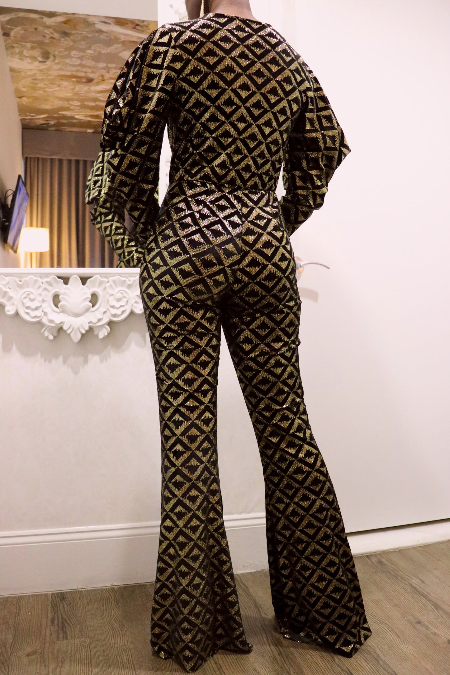 Back view of Model wearing a geometric-patterned jumpsuit standing in a room with a mirror and lamp.