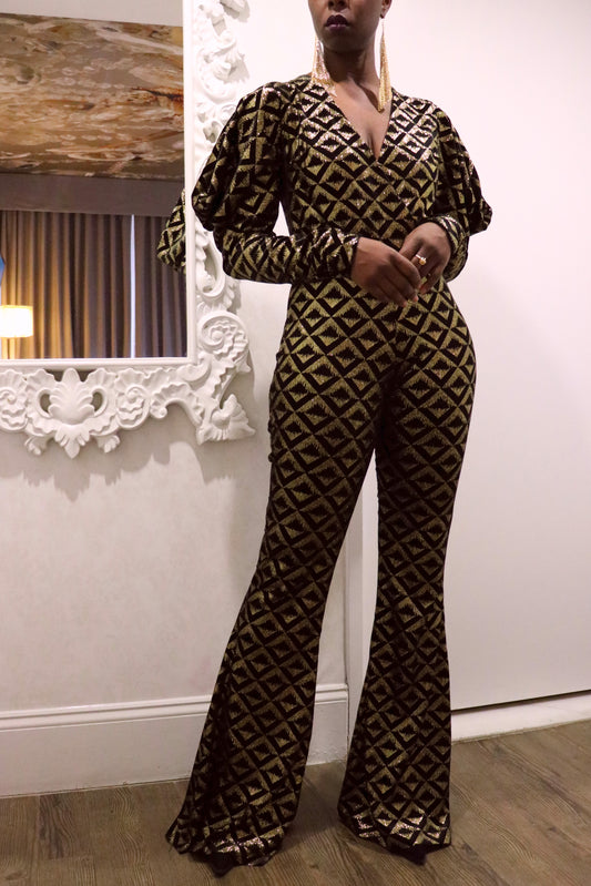 Model wearing a black and gold geometric-patterned jumpsuit standing in a room with decorative elements.