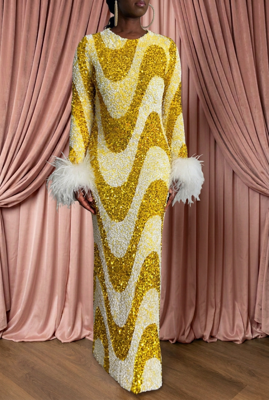 A woman wearing a long-sleeve maxi dress covered in gold and white sequins in a wavy pattern, with white feather trim at the cuffs, standing indoors against white paneled doors.
